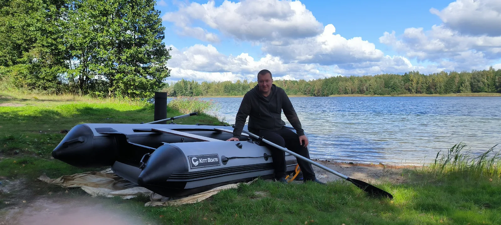 Владелец с веслом стоит рядом с тёмной лодкой KITT BOATS у озера