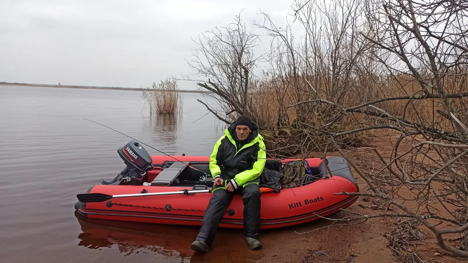 Рыбак в тёплой экипировке сидит в красной лодке KITT BOATS у берега